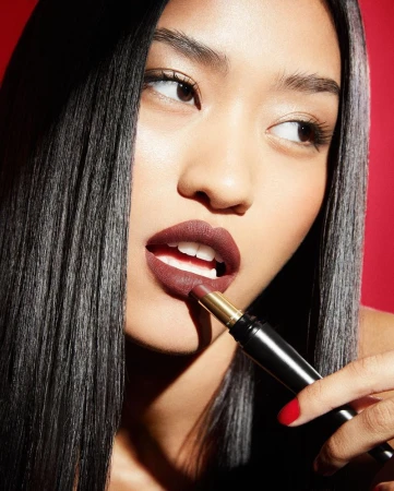 Close-up portrait of a woman applying dark lipstick with a stiletto lipstick tube against a bold red background.