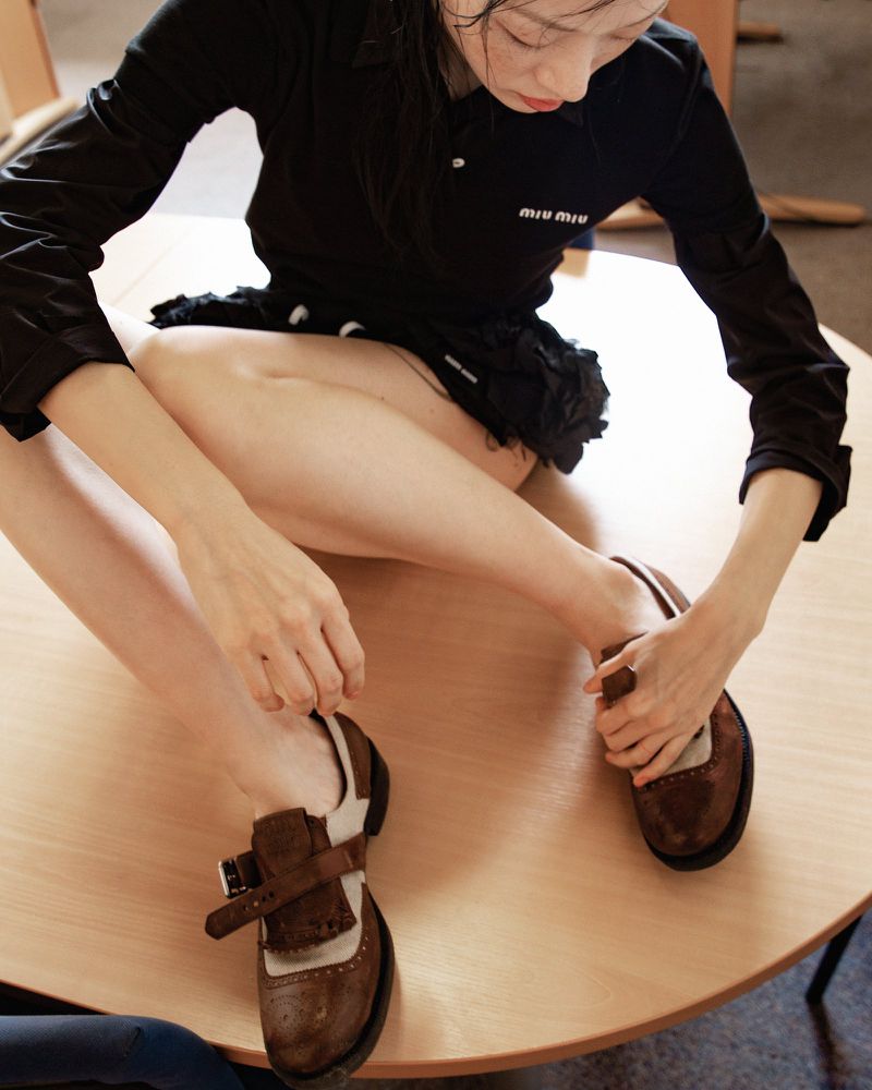 Seated woman in a black top and frilly skirt fastens a brown Mary Jane shoe at a round wooden table with light coming from above.