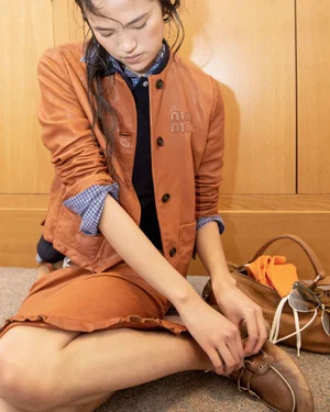 Model tying boat shoes wearing a tan leather jacket and skirt with a handbag nearby against wood panels.