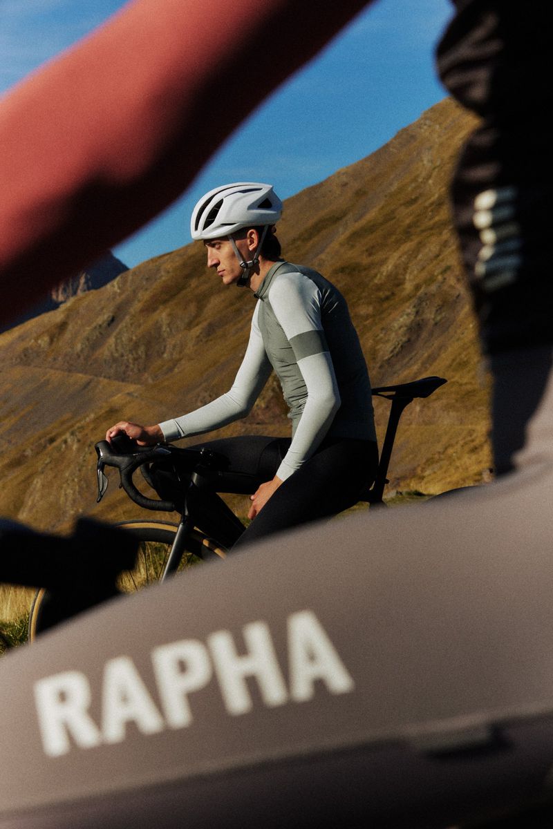 Cyclist wearing a white helmet and grey jersey riding on a bicycle on a sunny day in front of a mountain landscape.