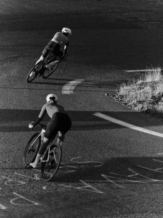 Two cyclists in helmets and sportswear ride racing bikes on an asphalt road, captured from an elevated perspective in monochrome.