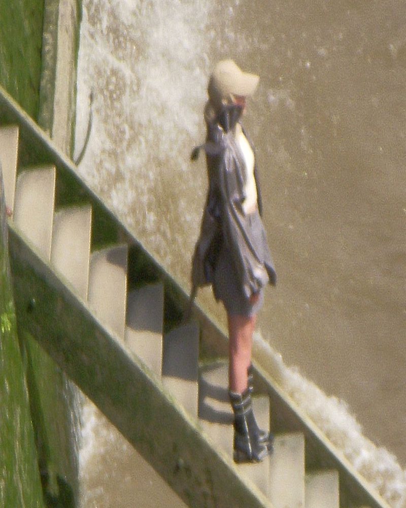 A woman with a beige hat and gray raincoat walks down wooden stairs alongside a waterfall, standing out against the white water.