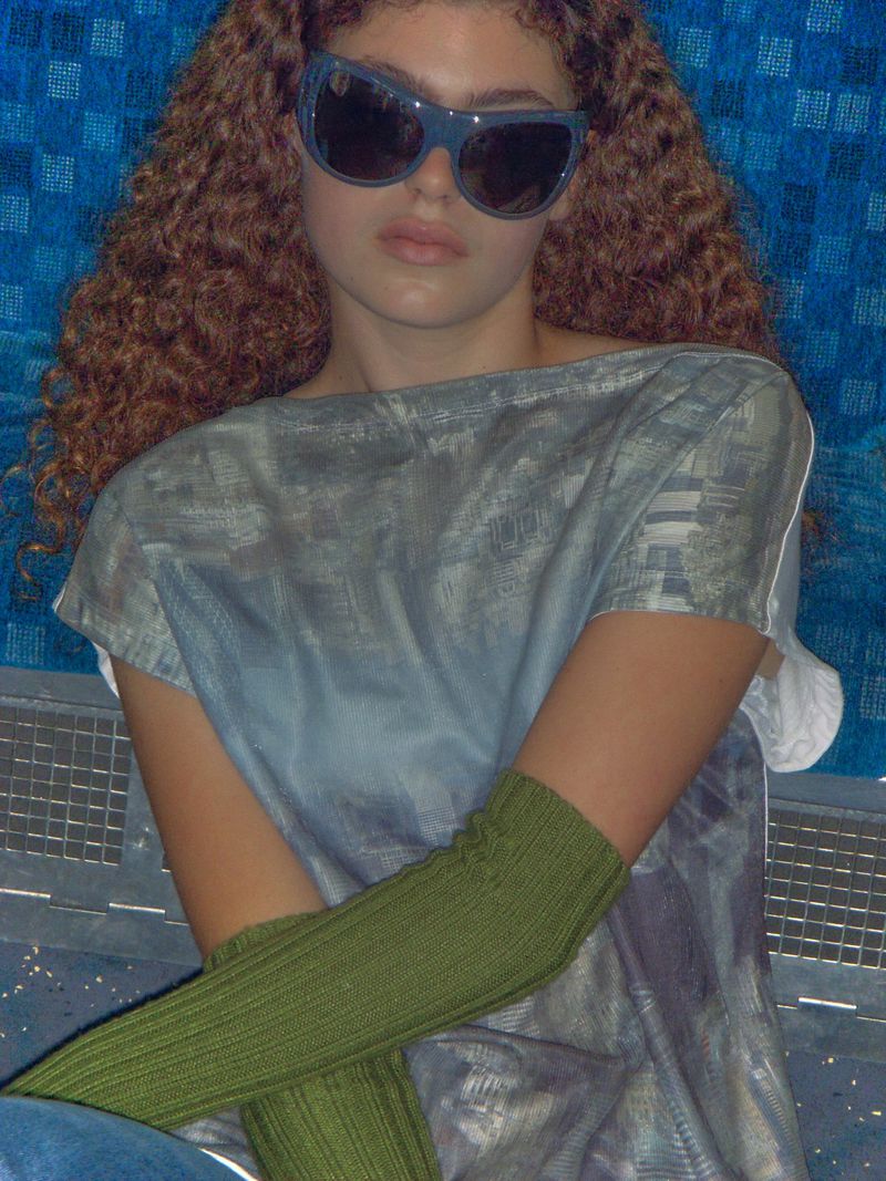 A young woman with curly hair wears sunglasses and olive green sleeves in an indoor setting with a blue patterned background.