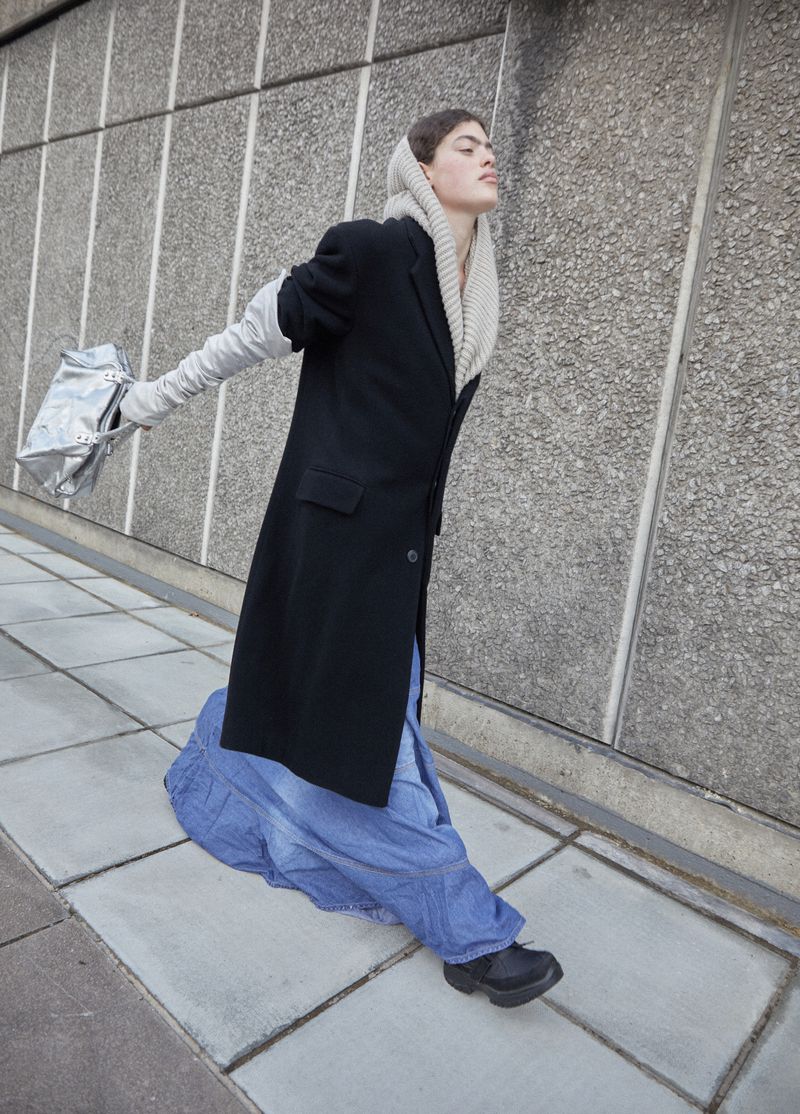 A fashion image showcases an adult model outdoors. The model wears a long black coat, long light gray gloves, and a long, full blue skirt.