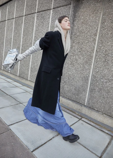 A fashion image showcases an adult model outdoors. The model wears a long black coat, long light gray gloves, and a long, full blue skirt.