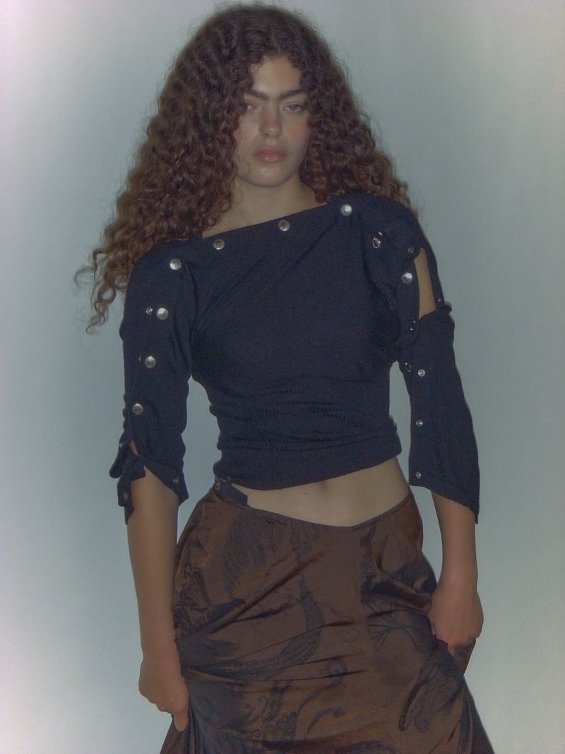 Young adult woman with curly brown hair poses in an indoor setting, wearing a dark top with metal accents and a brown skirt.