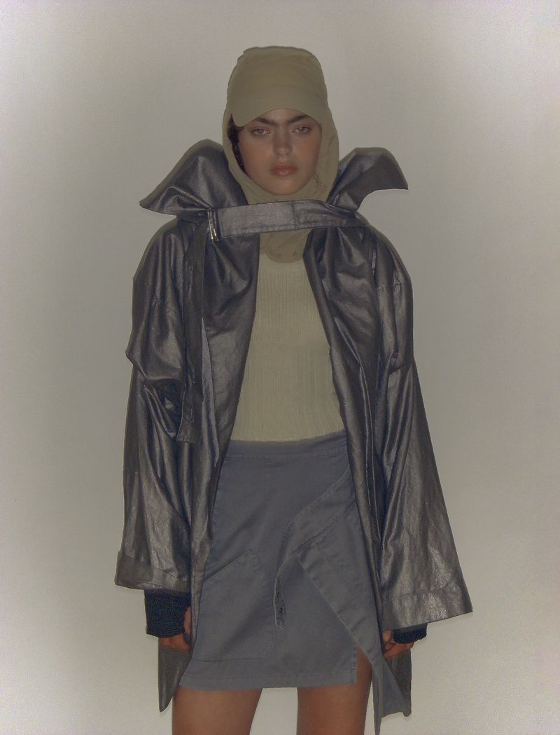 Woman poses in a metallic coat, grey skirt, and beige head covering against a light grey backdrop in a studio fashion shoot.