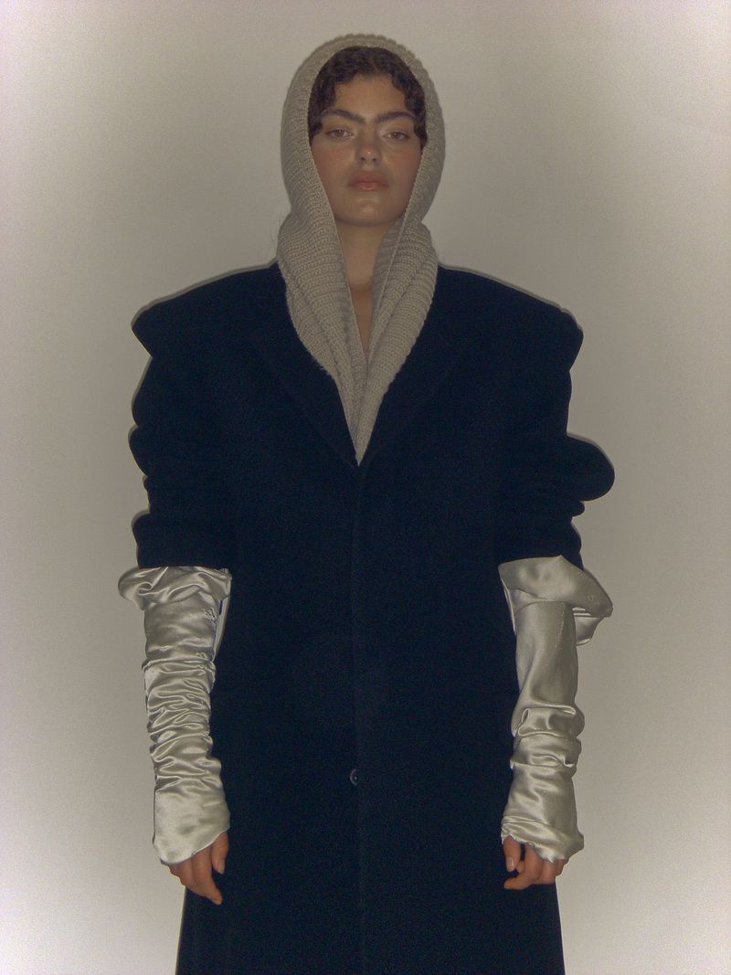 Portrait of a woman wearing a dark coat and a light-colored knitted hood with long satin sleeves against a neutral background.