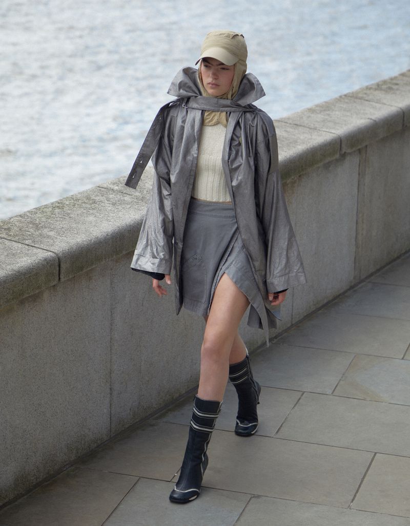 A woman walks along a stone walkway in a silver jacket, gray skirt, and black boots with a body of water in the background.