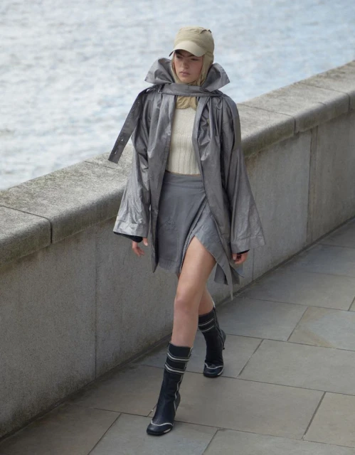 A woman walks along a stone walkway in a silver jacket, gray skirt, and black boots with a body of water in the background.