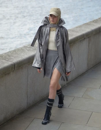 A woman walks along a stone walkway in a silver jacket, gray skirt, and black boots with a body of water in the background.