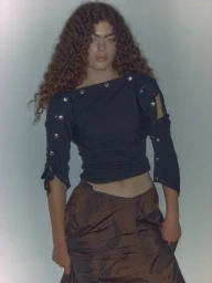 Model Isla Gaskin posing with long curly hair in a black buttoned cutout top and brown patterned skirt in front of a studio backdrop.