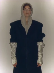 Model Isla Gaskin wearing a dark structured coat, a grey knit hood, and long ruched satin gloves in a studio portrait.