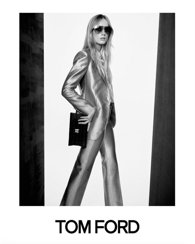 A blonde model presents a metallic suit and black purse for Tom Ford in a studio setting, standing against white and dark backgrounds.