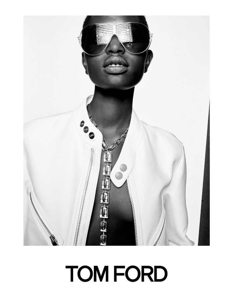 Black-and-white portrait of an adult female wearing aviator sunglasses and a white jacket, with a long pendant necklace.