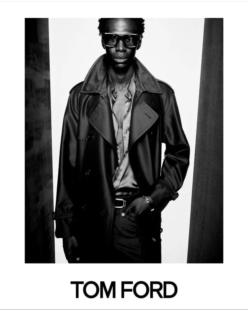 A Black male model wearing dark oversized sunglasses, an unbuttoned shirt, and a belted trench coat for a Tom Ford campaign.