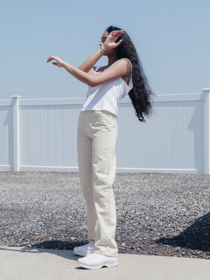 Young woman with long, dark curly hair shielding eyes in bright sunlight, wearing white tank top and beige pants, standing outdoors.