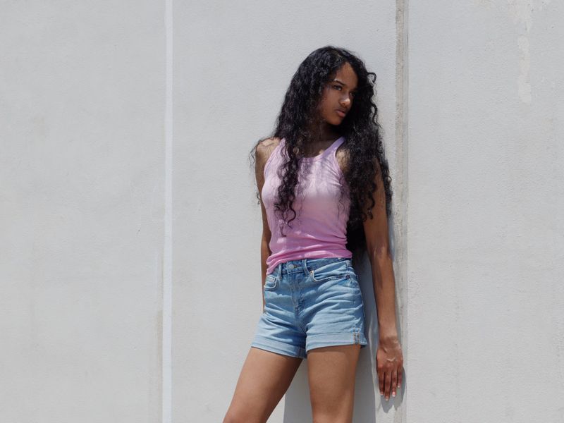 Woman with long dark curly hair leaning against a pale concrete wall wearing a pink tank top and cuffed denim shorts.