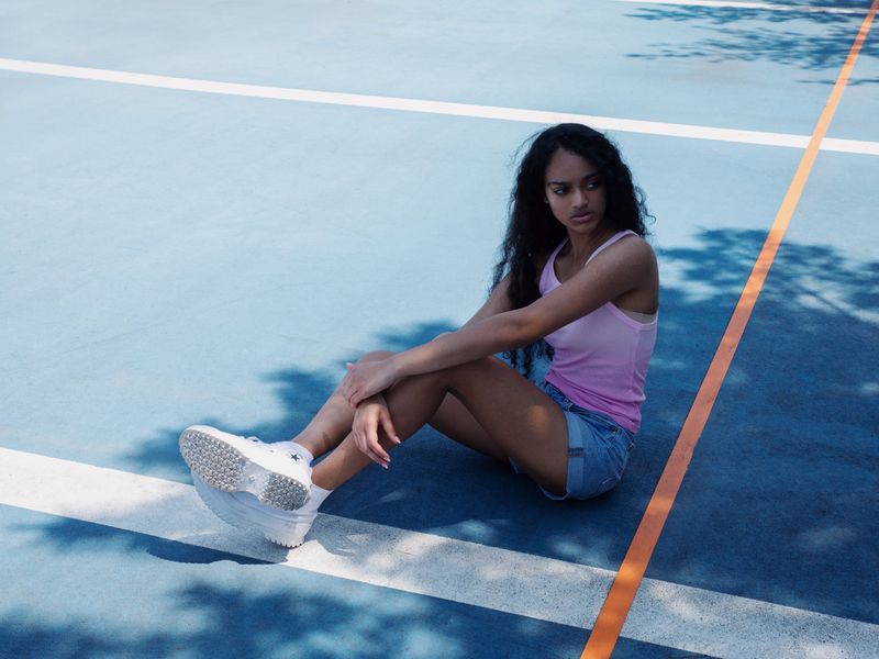 Young woman with long dark hair sits on a blue court with white lines, wearing a pink tank top, jean shorts, and white sneakers.