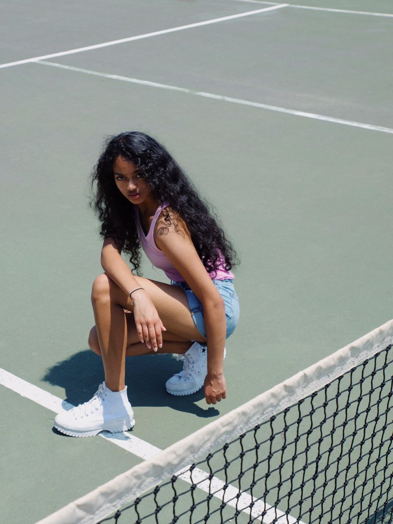 Young woman with dark curly hair, wearing a pink tank top and denim shorts, poses on a sunny tennis court by the net.