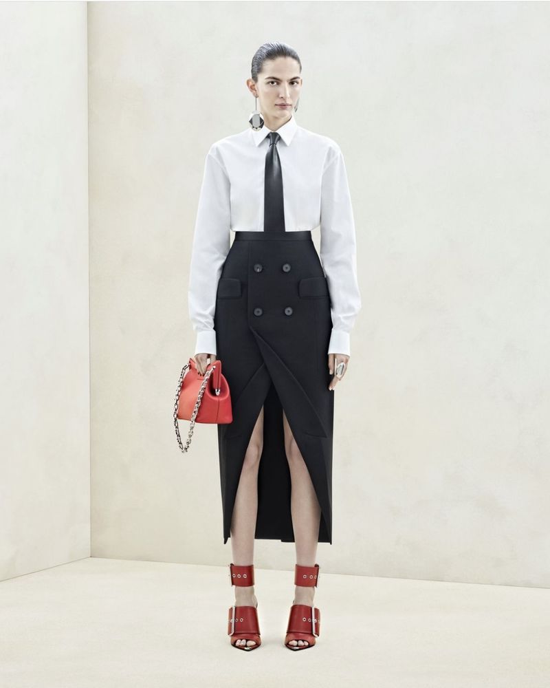 Fashion editorial shot of an adult female model posing in a white shirt, black skirt, red heels and purse in a photo studio.