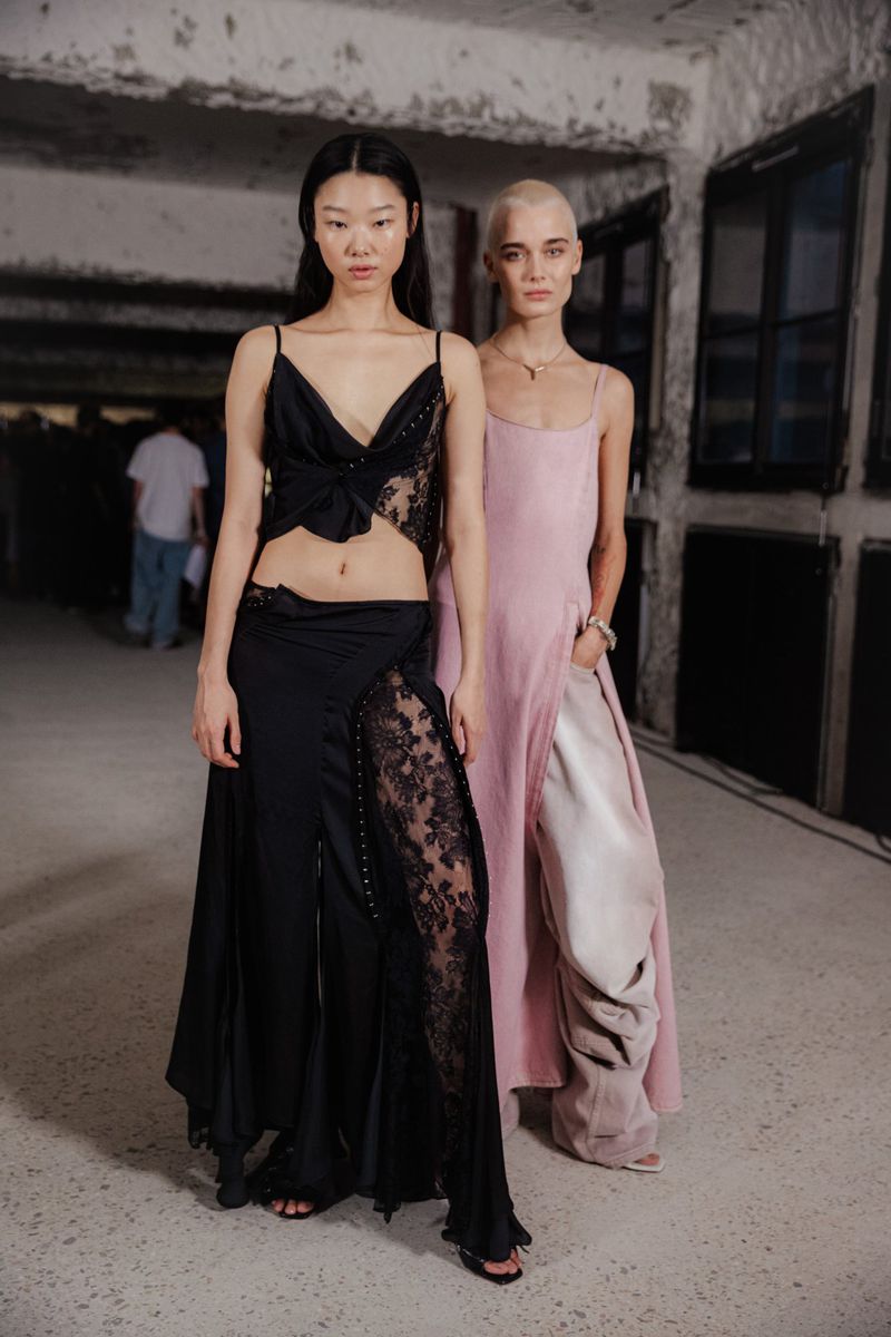 Two women pose in a textured hallway, one in a black lace dress, the other in a pink gown, in a fashion forward editorial.