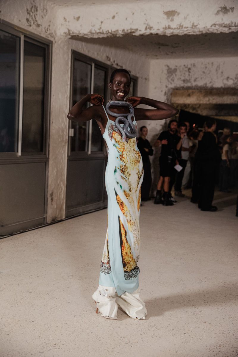 Backstage shot of an adult Black female model in a light blue dress with snake necklace posing during a fashion show indoors.