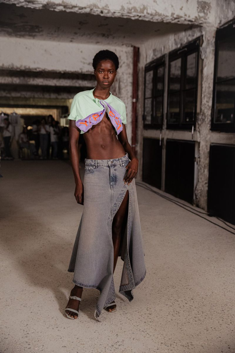 Runway fashion shot: A slender Black woman models a light green top and denim skirt with a slit, standing indoors on the runway.