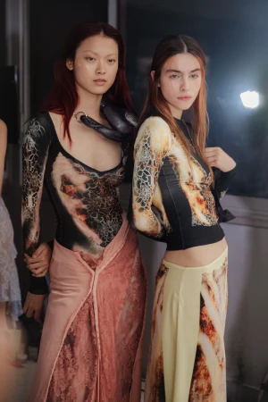 Two fashion models pose indoors wearing long-sleeved tops with abstract patterns and matching pastel maxi skirts in a studio setting.
