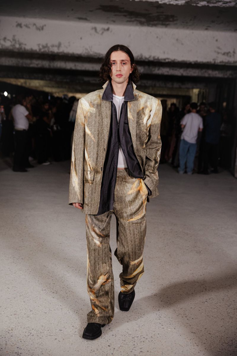 Runway fashion show featuring a male model in a patterned suit walking in an urban environment with blurred spectators in the background.