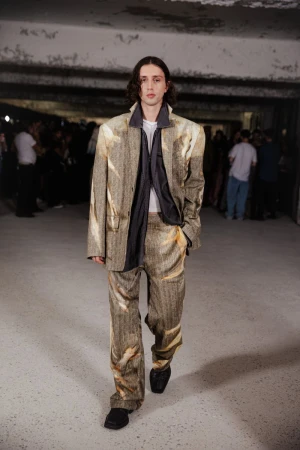 Runway fashion show featuring a male model in a patterned suit walking in an urban environment with blurred spectators in the background.