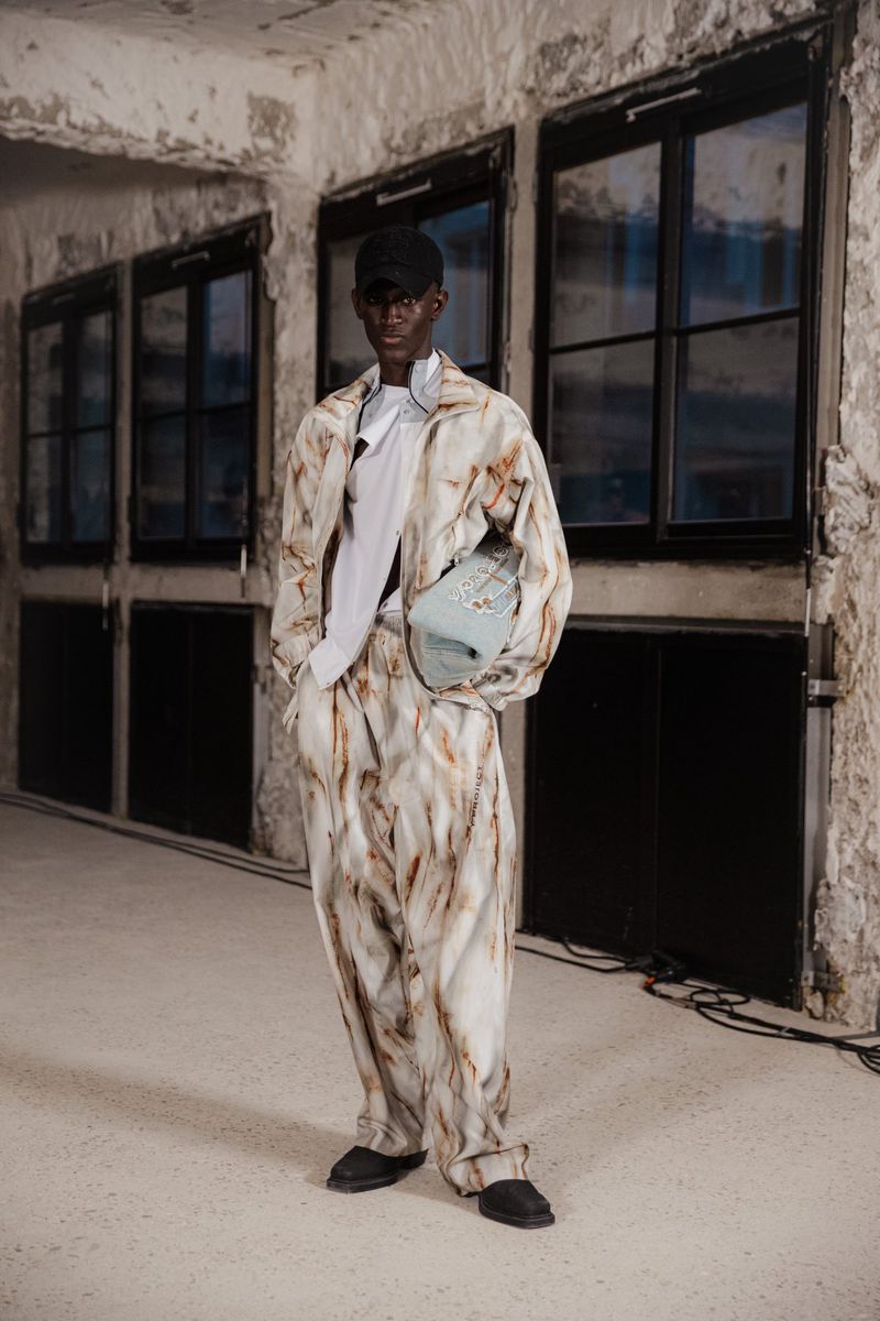 Man in marble-print jacket and trousers with baby blue bag in an industrial setting with weathered walls and dark-framed windows.
