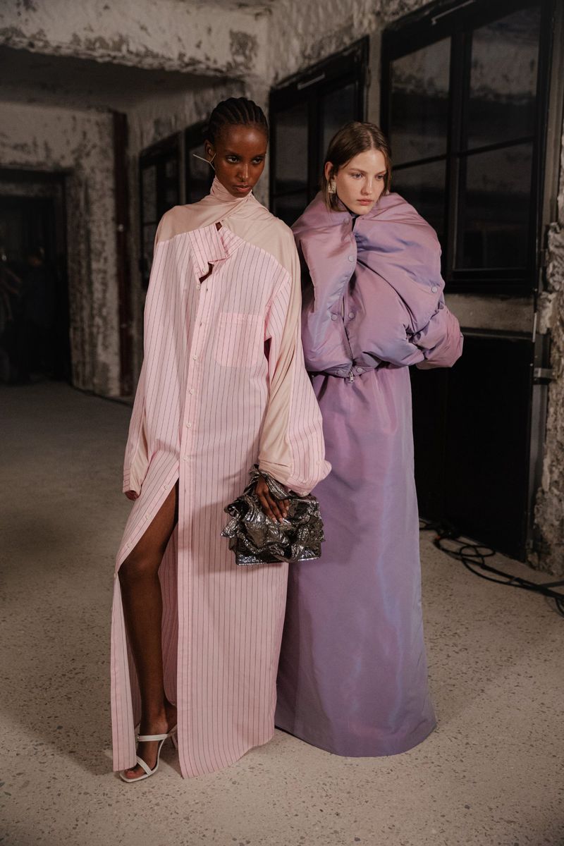 Two fashion models pose indoors. One wears a pink striped shirt dress with a silver bag, and the other wears a lavender puffer dress.