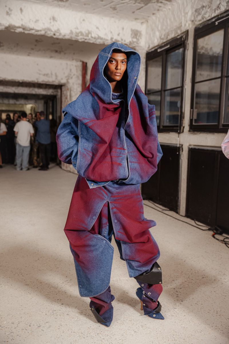 A model in an oversized blue and red denim outfit with a hood, stands in an indoor setting with windows and other people visible.