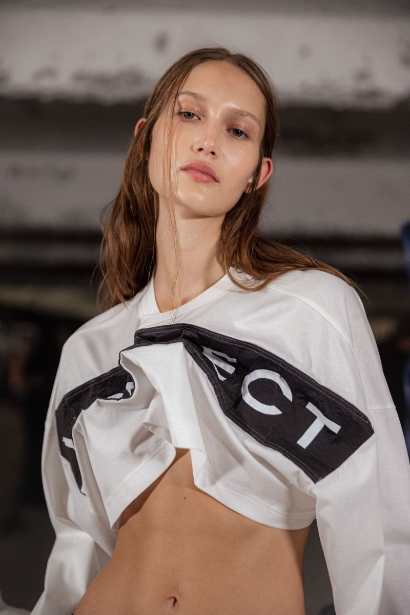 A young, fair-skinned woman poses in a white crop top with black text. Her medium-length brown hair is loose, with a neutral facial expression.