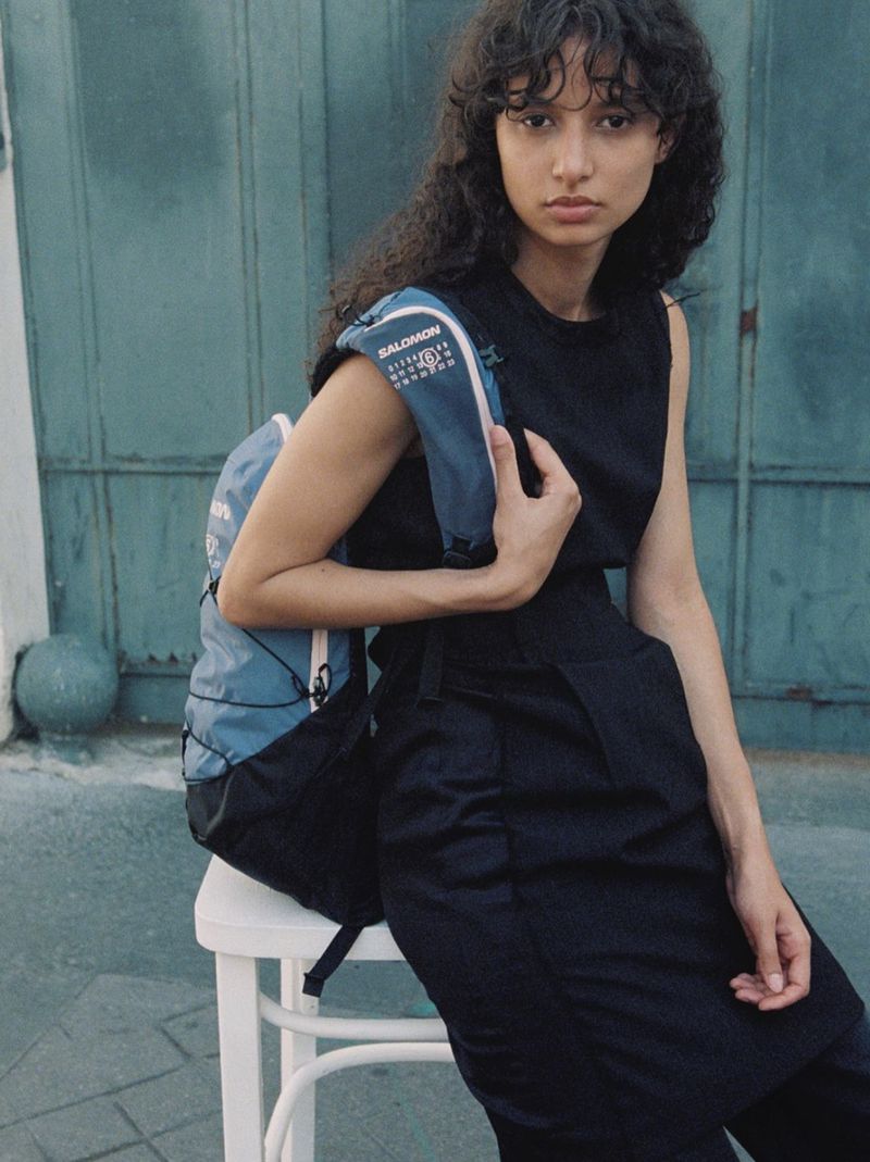 An adult female sits on a white stool, wearing a black outfit and a blue backpack against a textured turquoise wall.