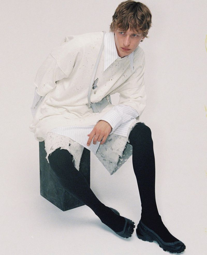 An adult male in casual fashion sits in a studio setting, wearing a white top, black tights, and dark shoes, with a minimalist backdrop.