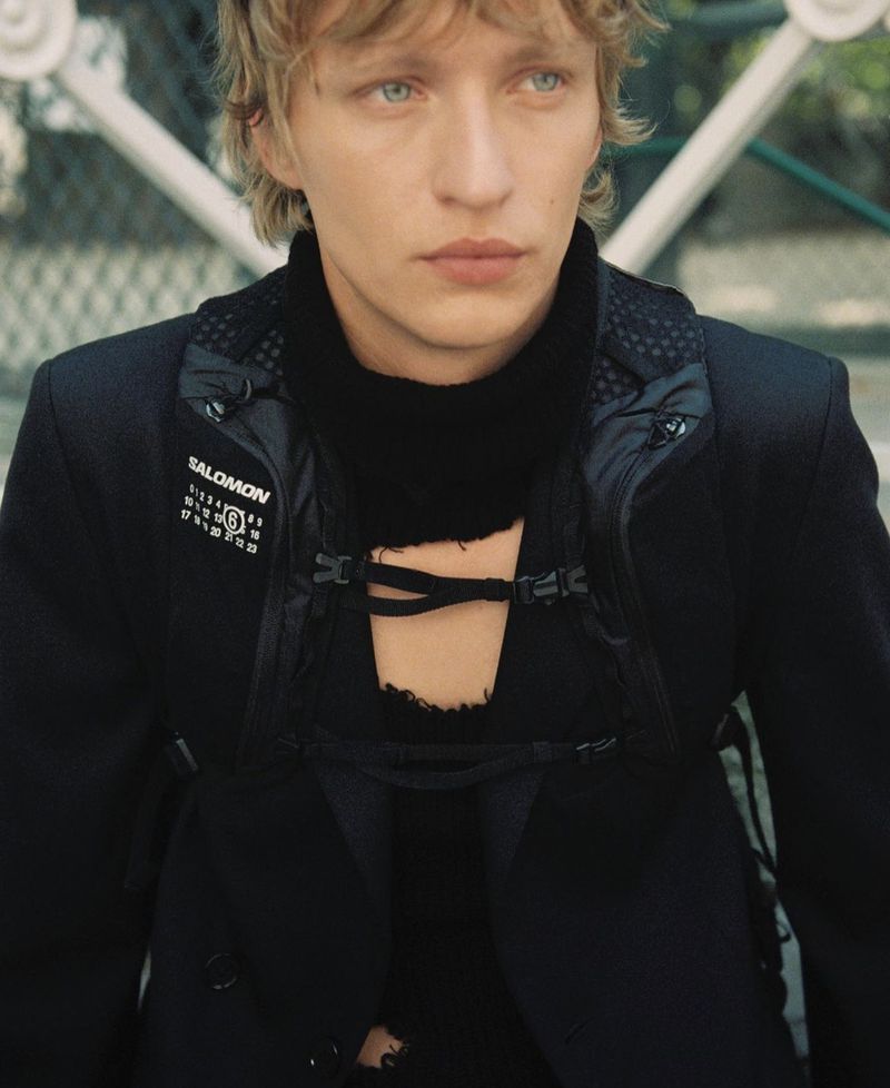 Close-up of a young male model with blond hair and blue eyes wearing a black jacket with Salomon logo, posing outdoors.