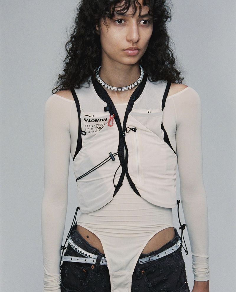 A woman poses in a studio wearing a light colored top, a white Salomon vest, and dark denim shorts against a grey backdrop.