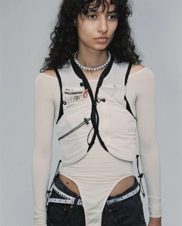Model with dark curly hair wearing an off-white tactical vest over a sheer top and dark denim jeans against a grey background.