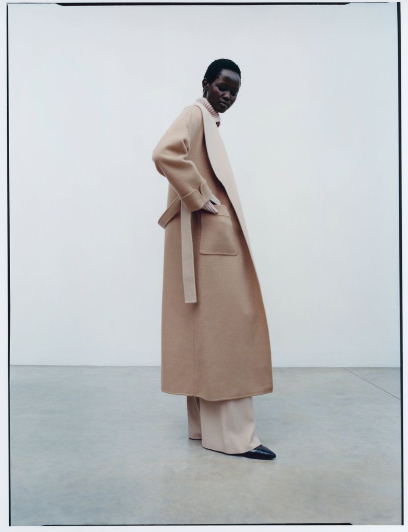 A Black female fashion model poses in an indoor photo studio wearing a beige wool coat, matching trousers and black shoes on a white background.