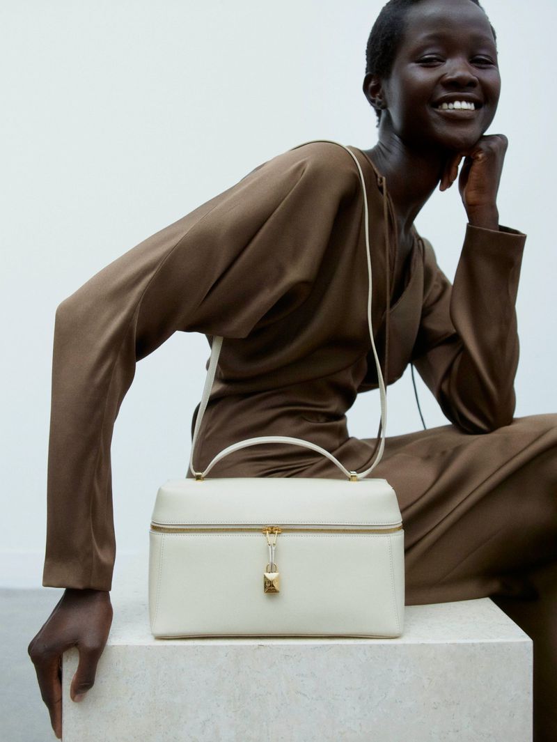 Woman in a brown dress sits next to a cream-colored handbag on a concrete block, smiling, studio lighting, and simple white backdrop.