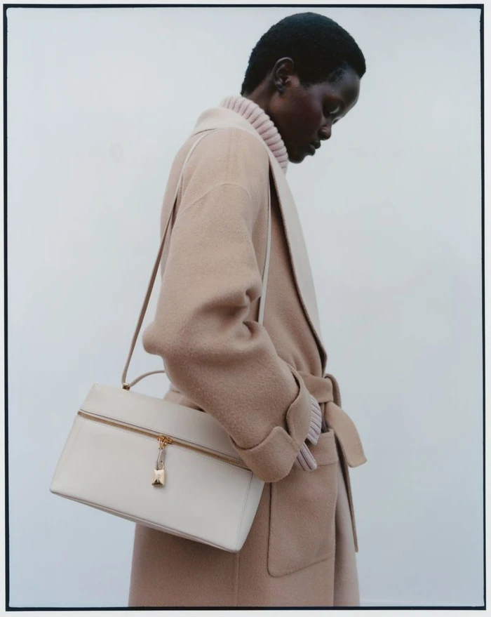 Model Anyiel Majok in profile wearing a camel belted wool coat and holding an off-white leather handbag against a pale studio backdrop.