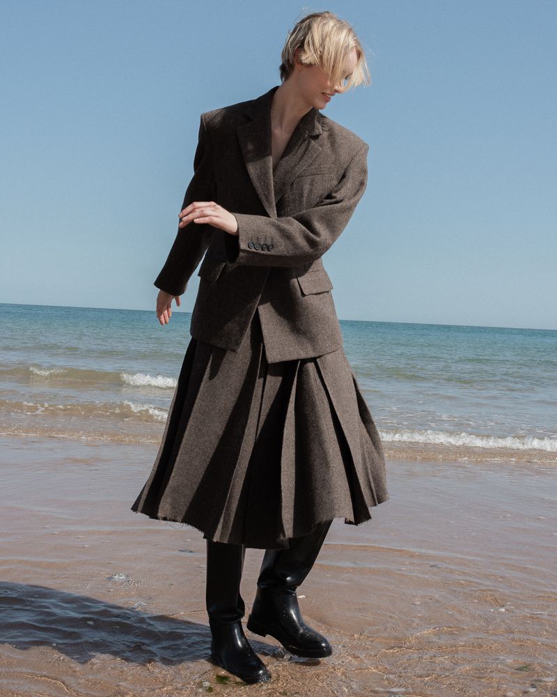 Model wearing a tailored brown blazer, pleated midi skirt, and black leather boots walking along a sandy beach by the water.