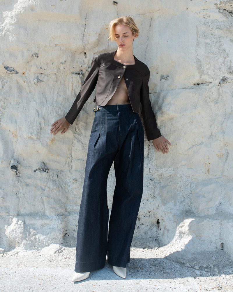 A stylish fair-skinned woman poses in a brown jacket and wide-leg jeans against a textured white wall, complemented by light-colored shoes.