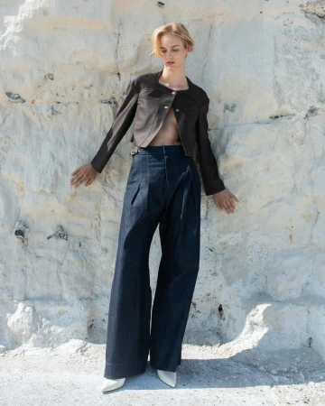 A stylish fair-skinned woman poses in a brown jacket and wide-leg jeans against a textured white wall, complemented by light-colored shoes.