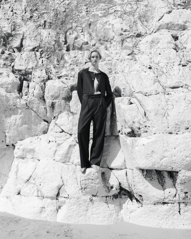 Monochrome shot of a woman in an open dark blazer and wide-legged pants standing on a rocky outcrop, against a craggy rock face.