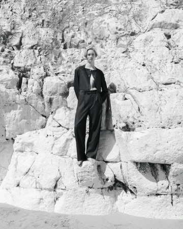 Monochrome shot of a woman in an open dark blazer and wide-legged pants standing on a rocky outcrop, against a craggy rock face.