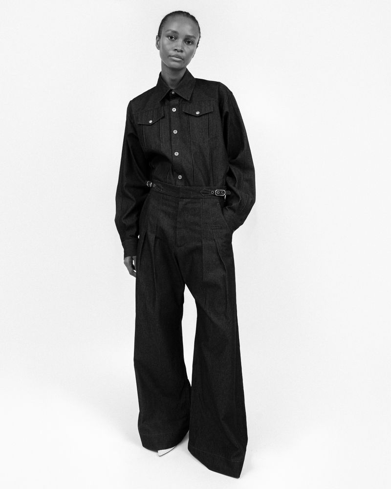 Monochromatic portrait of an adult woman wearing a buttoned dark denim shirt and wide-leg trousers in a fashion studio setting.