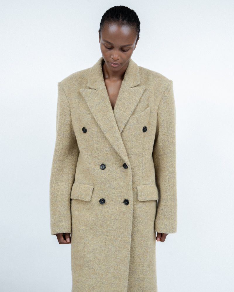 Fashionable woman in a tan double-breasted overcoat against a white background in a studio setting, looking down in a medium shot.